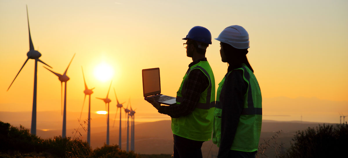 Zwei Arbeiter mit einem Laptop vor Windmühlen im Sonnenuntergang