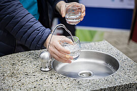 Frauenhände, die am Trinkbrunnen 2 Gläser mit Wasser füllen.