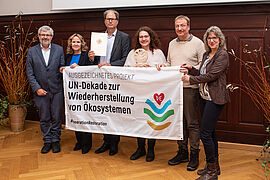 Steffi Lemke hält mit fünf anderen ein Banner mit der Aufschrift "Ausgezeichnetes Projekt: UN-Dekade zur Wiederherstellung von Ökosystemen"