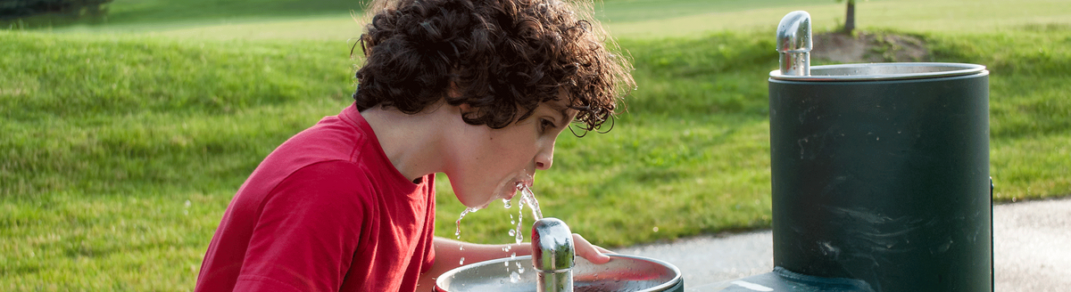 Junge mit lockigem Haar, der aus einem Wasserbrunnen in einem Park trinkt
