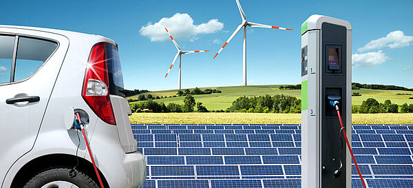 Elektroauto beim Auftanken an einer Ladestation, im Hintergrund eine Solaranlage und Windkraft-Anlagen