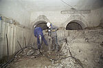 Zwei Arbeiter stemmen mit Presslufthämmer den Boden in einem unterirdischen Raum auf.