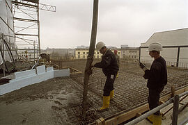 Zwei Arbeiter mit Helmen gießen auf einem großem Gebäude einen Teil der Decke mit Beton aus. Im Hintergrund die obersten Etagen umliegender Häuser.