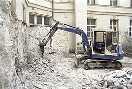 In einem Innenhof werden mittels eines Presslufthammers an einem Minibagger Reste eines Anbaus von einer Gebäudewand entfernt.