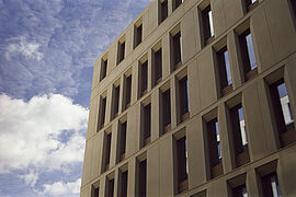 Außenansicht der sandsteinfarbenen Fassade des Neubaus, die von schmalen Holzfenstern geprägt ist; darüber blauer Himmel mit Wolken