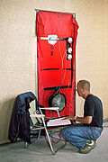 Ein Mann hockt vor einer mit Plastik abgeklebten Tür, in die ein Gebläse eingelassen ist. Auf seinem Schoß ein Block, auf dem er Messergebnisse von den angeschlossenen Geräten notiert.