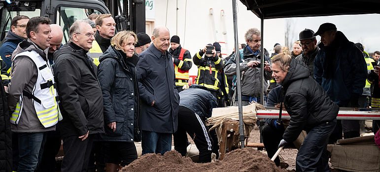 Steffi Lemke und Olaf Scholz schauen beim Sandsäcke füllen zu. 