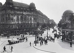 Historische Schwarzweißaufnahme eines Platzes mit Gleisen, Pferdefuhrwerken, Passanten und Fahrradfahrern, Litfaßsäule und imposanten Häuserfassaden und Bauten.