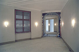 Ein Gang im Rohbauzustand mit leuchtenden Wandlampen einem Fenster, Türausschnitten und einem bogenförmigen Durchgang.