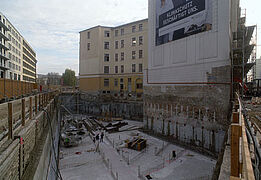 Die Baugrube des Neubaus mit gegossener Sohlenplatte, angrenzend und im Hintergrund andere Gebäude.