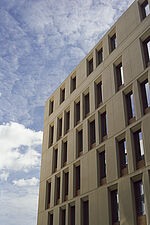 Außenansicht der sandsteinfarbenen Fassade des Neubaus, die von schmalen Holzfenstern geprägt ist; darüber blauer Himmel mit Wolken