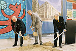 Drei Männer in Anzügen heben vor einem Berliner Mauerrest mit Schaufeln symbolisch eine kleine Grube aus, neben ihnen ein Pult mit Mikrofon.