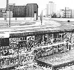 Schwarzweißaufnahme mit Stück bemalter Berliner Mauer im Vordergrund dahinter der Grenzstreifen, dann wieder eine Mauer mit Wachturm und jenseits derer Alt- und Neubauten.